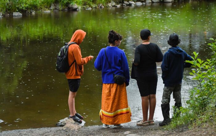 Four young people stand on the banks of a calm stream as the rain dimples its glassy green surface. One of the youth is wearing a bright orange ribbon skirt.