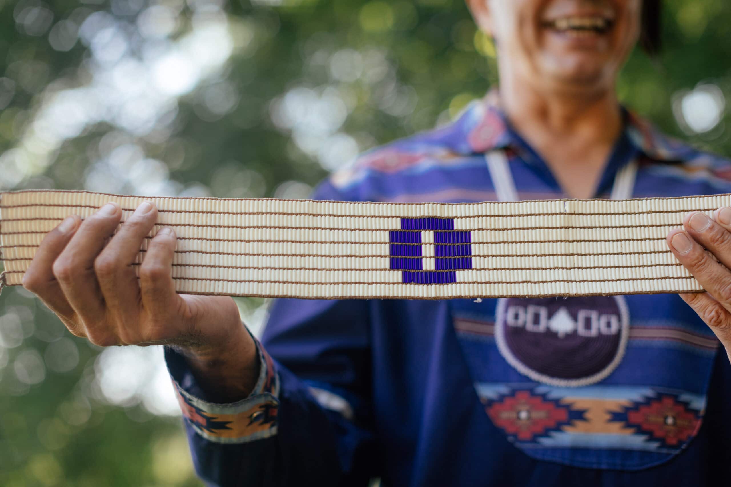 A laughing man holds up a beaded belt. This belt is white. In its centre is a purple circle containing a small rectangle of white. The man is wearing a beaded Hiawatha Belt medallion.
