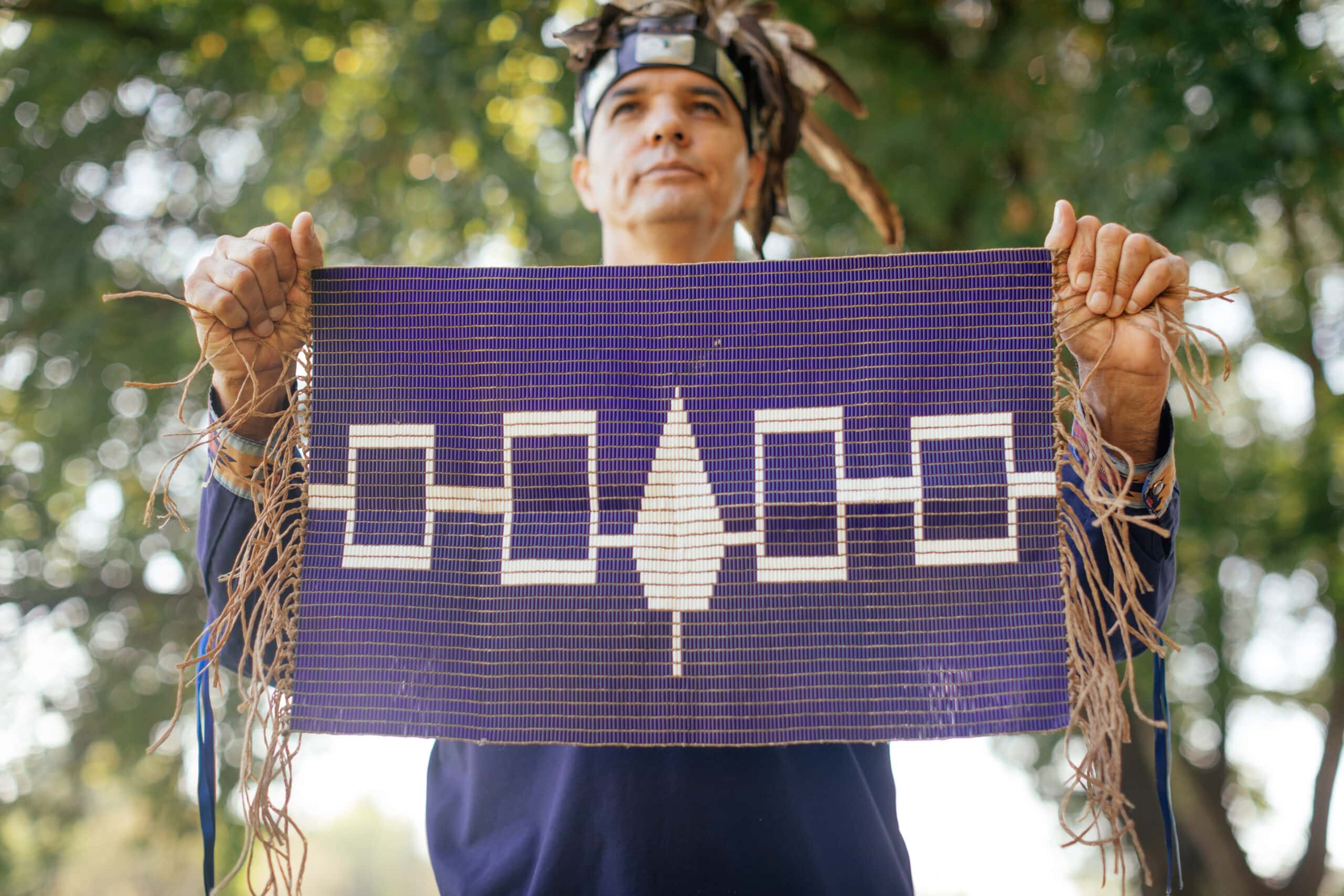A Cayuga man wearing a headdress holds up a purple beaded belt. On the belt are four white rectangles connected together with a white tree in the centre, which is also connected to the rectangles. There are trees in the background.
