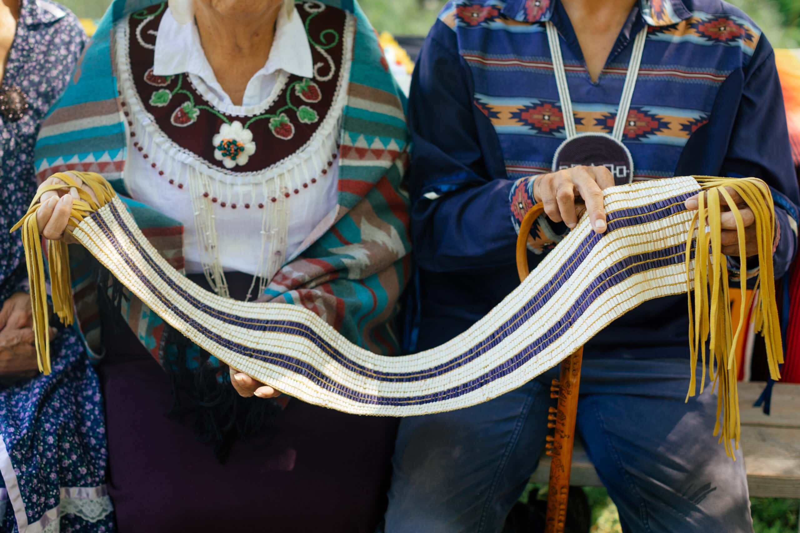 A Haundenosaunee woman and man between them hold a beaded belt which shows three stripes of white and two stripes of purple beads in the centre.