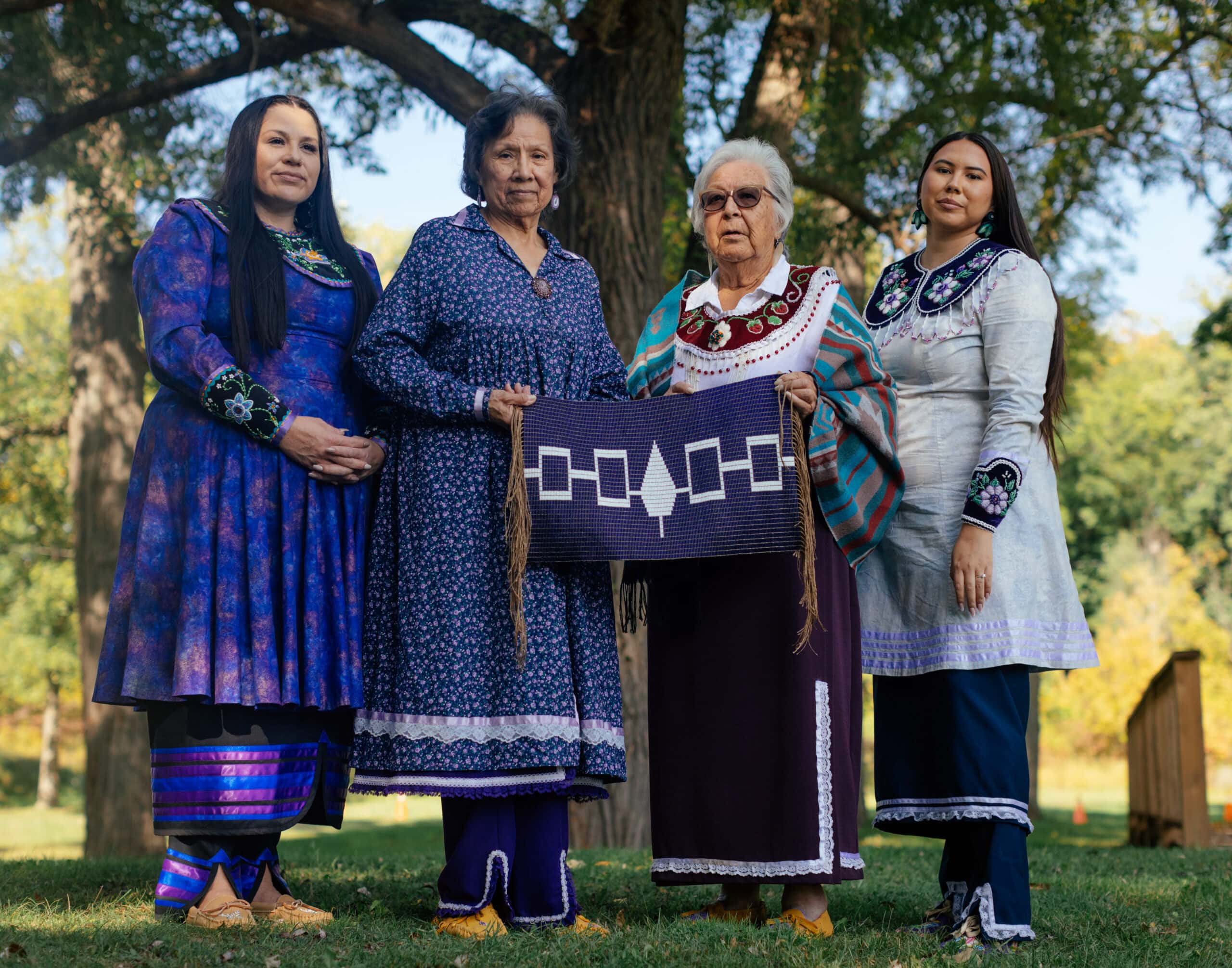 Four Haudenosaunee women of varying ages wearing purple and white hold a beaded belt. On the belt are four white rectangles connected together with a white tree in the centre, which is also connected to the rectangles.