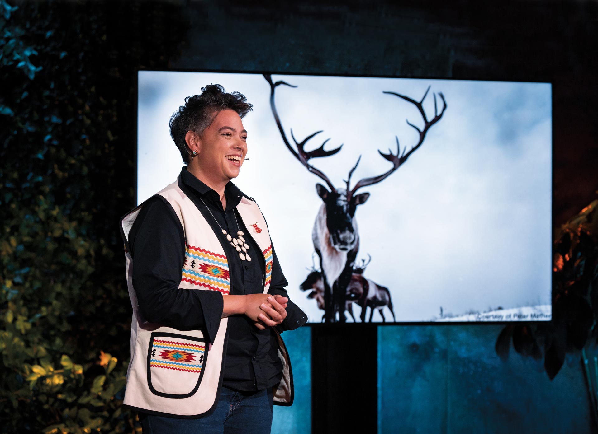 An Innu woman wearing a white patterned vest stands in front of a projector screen on a stage. On the screen is a photograph of a herd of caribou with long elaborate horns.