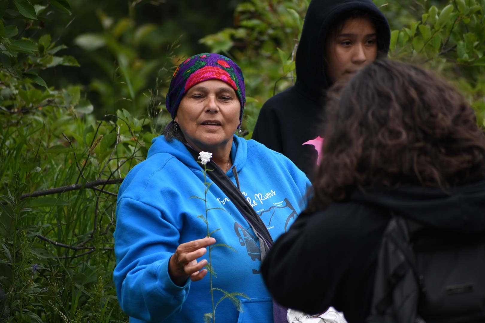 A woman wearing a blue hoodie and a purple kokum scarf holds a slender white flower. Standing next to her are two youth.