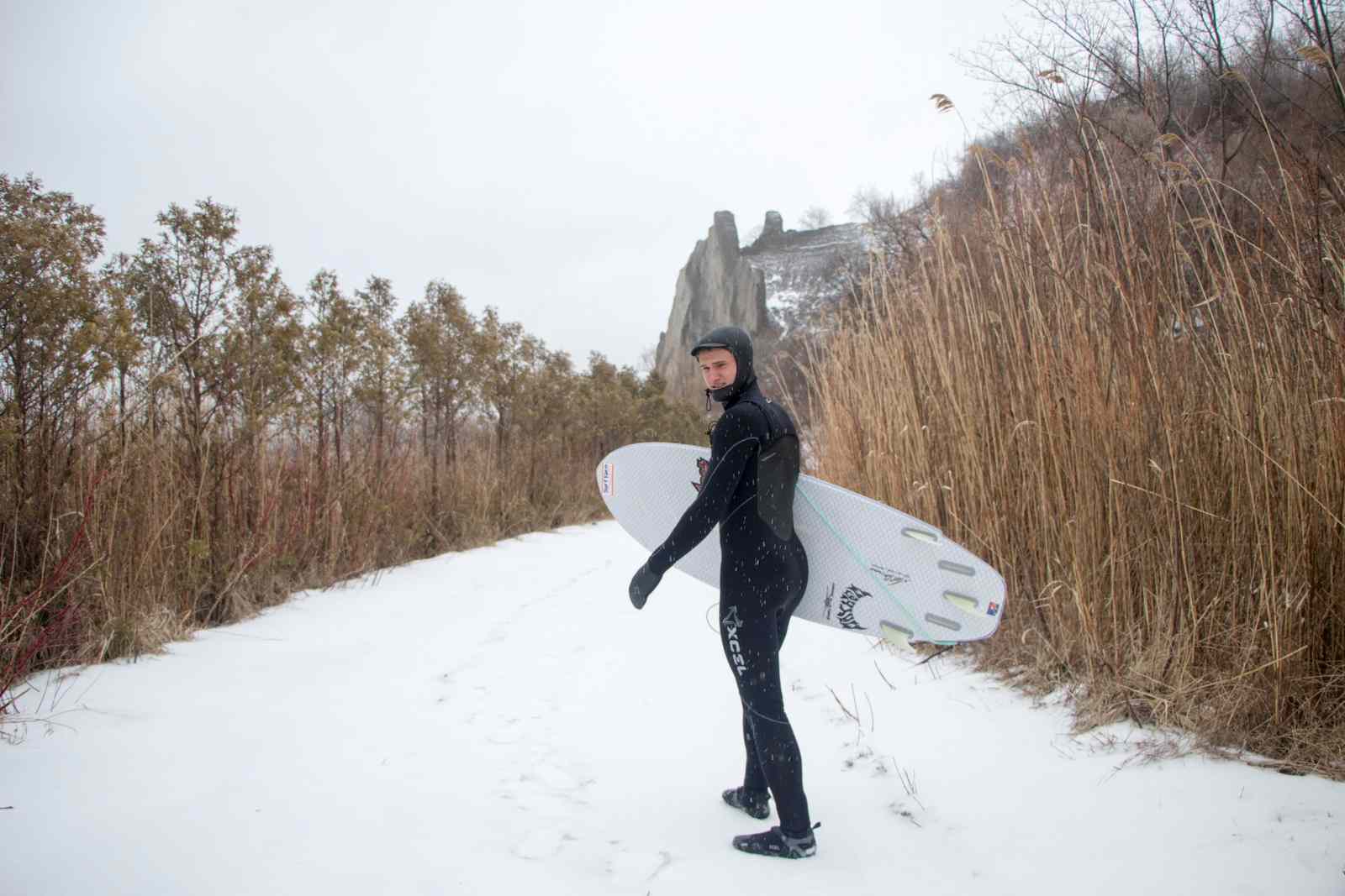 Man in a wetsuit carries a surfboard down a snowy path.