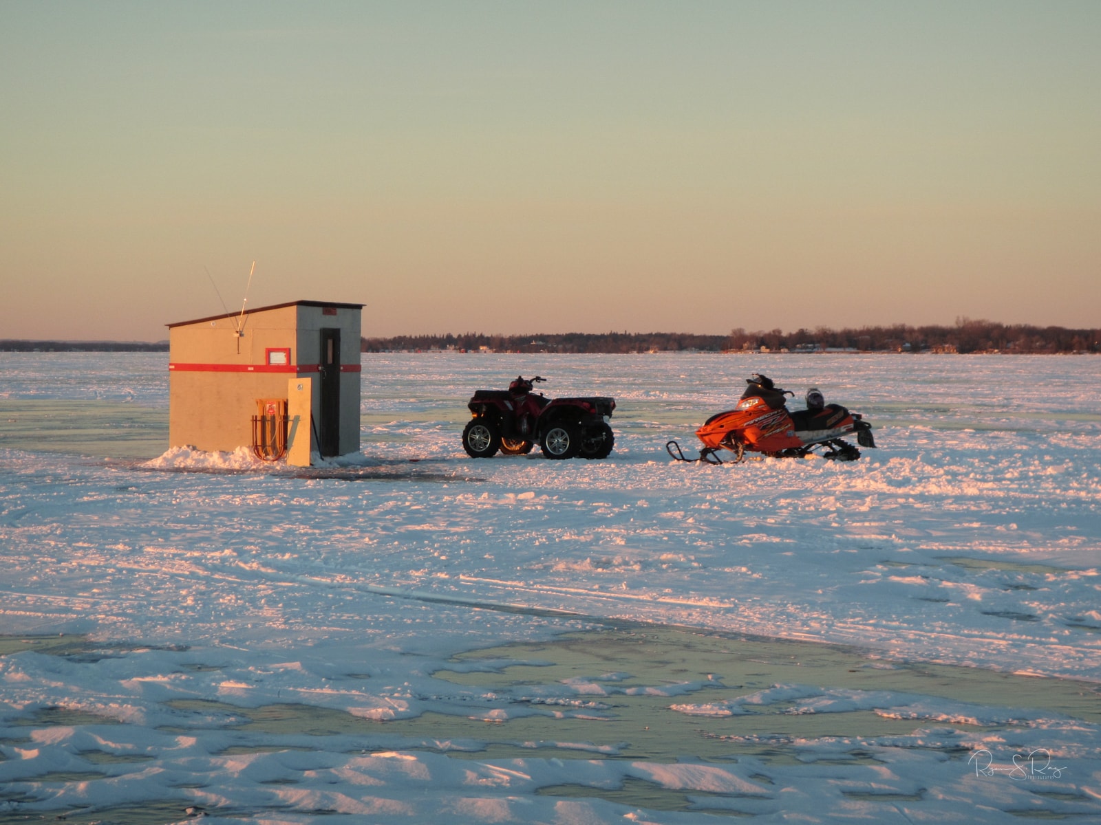 An ice fishing hut glows under the setting sun. Snow has accumulated on the lake and an ATV and snow mobile sit beside the hut.