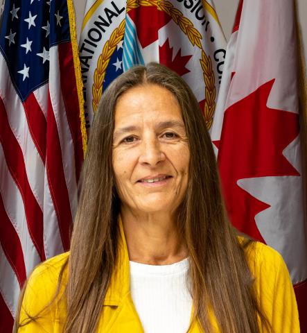 A portrait of Dr. Susan Chiblow in a sunny yellow blazer. Behind her are the flags of the USA, the IJC, and Canada.