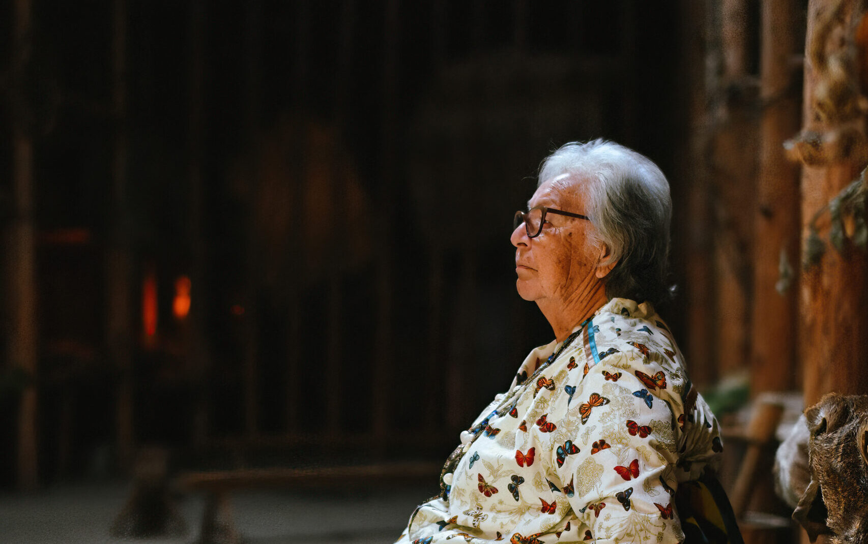 Side profile of a Cayuga Elder in traditional women's clothing sitting in a dark longhouse